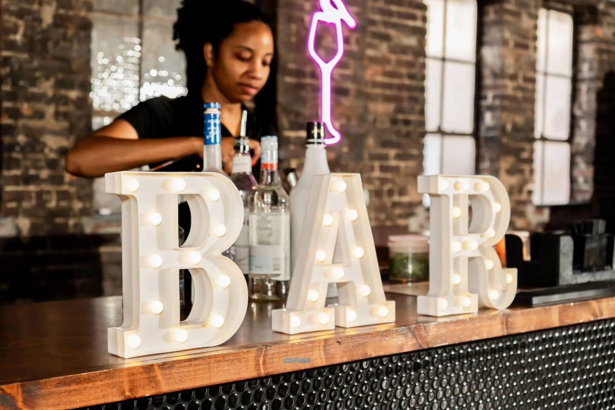 Large illuminated marquee letters spelling BAR on a wooden bar counter in a bar setting, with a bartender visible in the background.